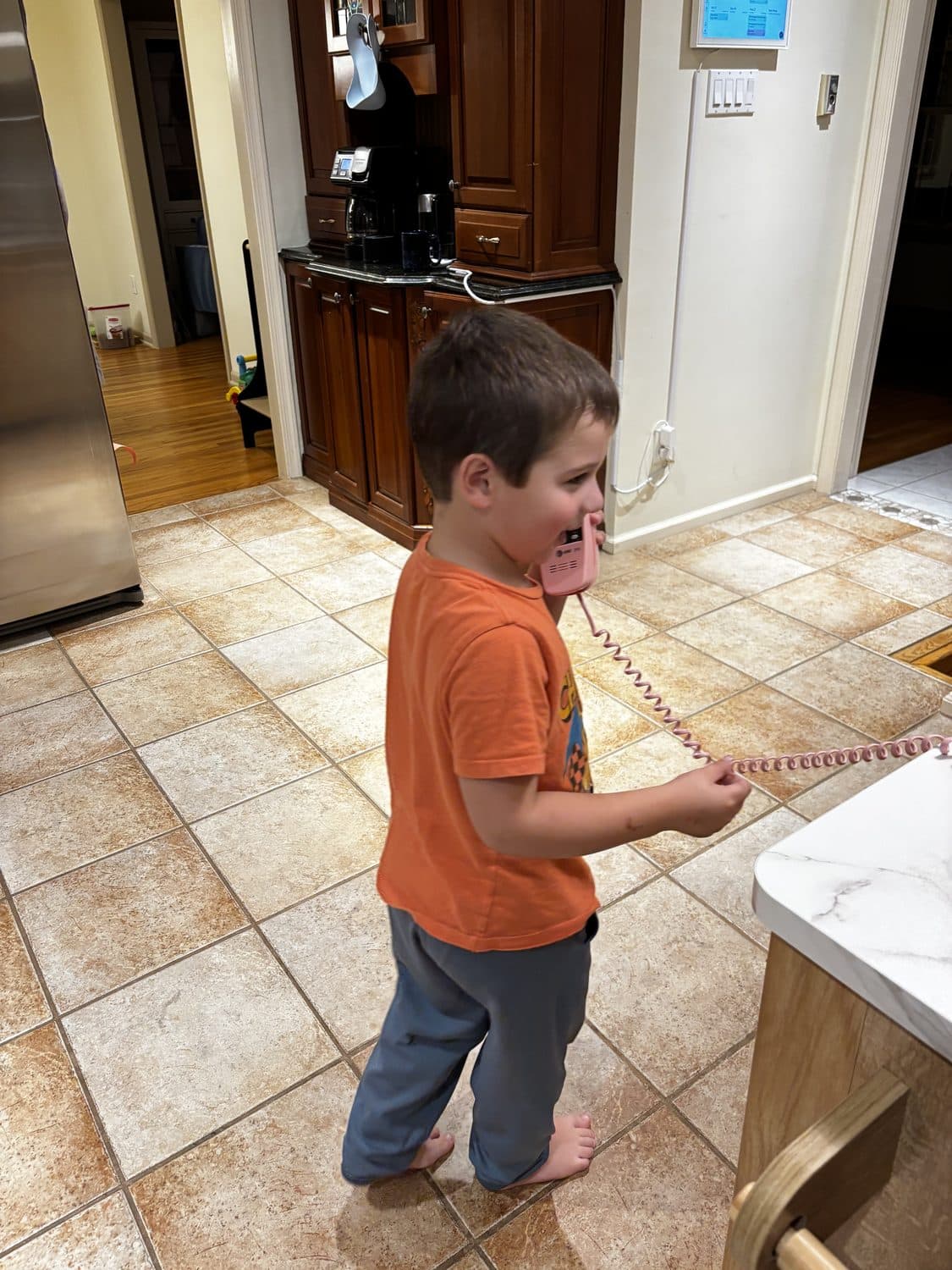 A young boy standing in a kitchen, talking into a pink corded handset with a coiled cord